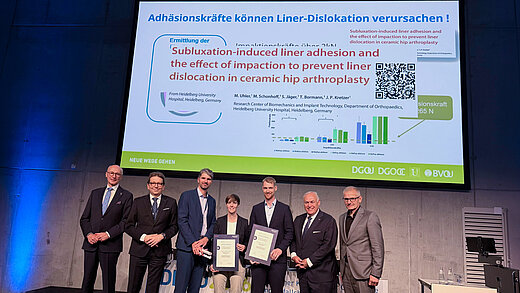 Award recipients and presenters on stage at DKOU 2026 beneath a slide on ceramic hip arthroplasty liner adhesion.